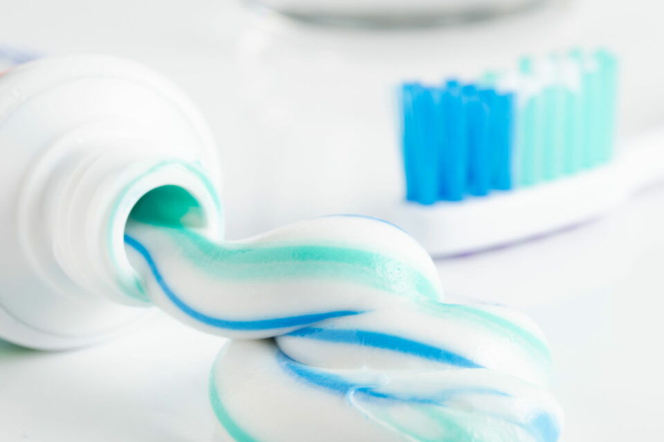 An open tube of toothpaste and a toothbrush lie on a washstand. Blue and white toothpaste oozes out of the tube